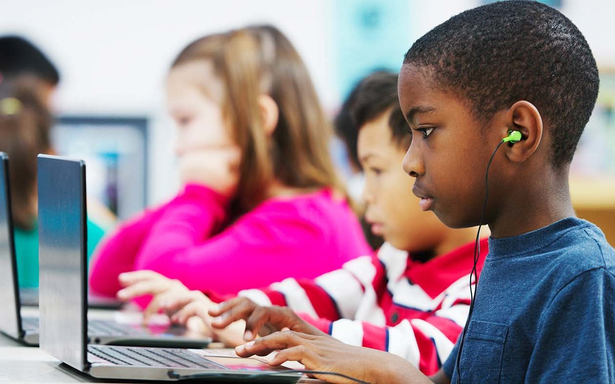 A multi-ethnic group of elementary age children are in the computer lab using laptops. A little boy is watching a video and is listening to music.